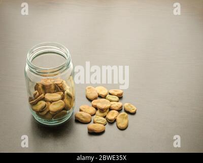 Fèves (Vicia faba) dans un bocal en verre sur la table en bois. Les fèves dans un plat en verre. Fond de bois. Les fèves sont déversées sur la surface en bois Banque D'Images