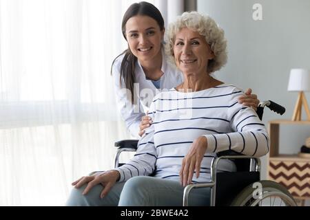 Une infirmière soignante encadre une femme handicapée âgée en fauteuil roulant, posant à l'intérieur Banque D'Images