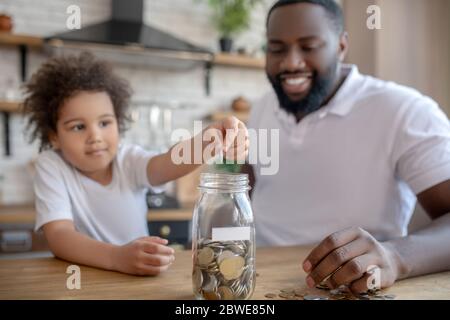 Mignon petit enfant aux cheveux bouclés mettant des pièces dans la banque de moulus Banque D'Images