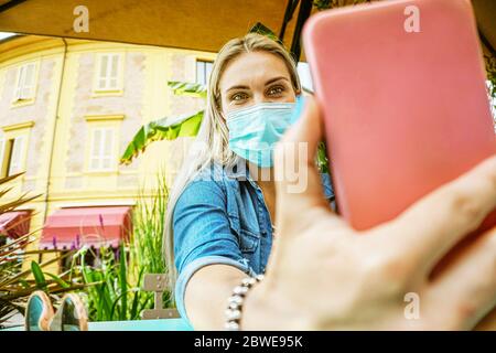 Fille dans un bar de jardin prenant un selfie avec son masque de visage pour être protégé du coronavirus - étudiant tenant smartphone dans un videocall dehors moi Banque D'Images