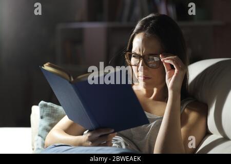 Femme avec problème de vue portant des lunettes lit livre la nuit à la maison Banque D'Images