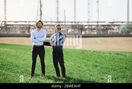 Deux jeunes ingénieurs se tenant à l'extérieur près d'une raffinerie de pétrole, discutant des questions. Banque D'Images