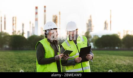 Deux jeunes ingénieurs avec une tablette à l'extérieur près d'une raffinerie de pétrole, discutant des problèmes. Banque D'Images