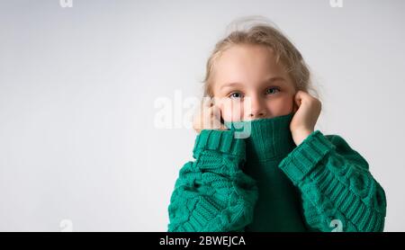 Adorable petite fille cachant la partie inférieure de son visage sous un col épais de pull chaud tricoté. Gros plan studio isolé sur espace de copie blanc Banque D'Images