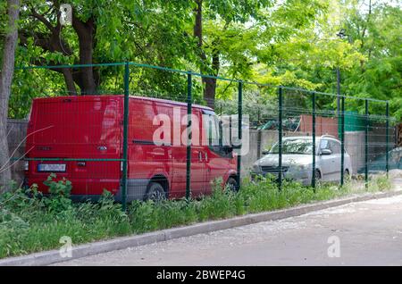 Nikolaev, Ukraine - 31 mai 2020 : parking clôturé avec maillage métallique sur la pelouse avant. Minibus rouge et berline grise à l'ombre des arbres devant un cdm Banque D'Images