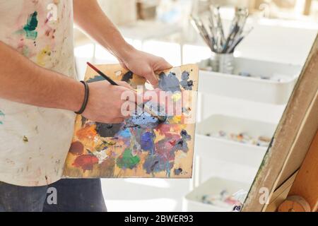 Gros plan d'un artiste masculin méconnu tenant une palette et mélangeant de la peinture tout en peignant une photo dans un studio d'art, espace de copie Banque D'Images