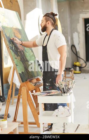 Portrait vertical complet d'un artiste contemporain barbu qui peint une image sur chevalet tout en travaillant dans un studio d'art éclairé par la lumière du soleil Banque D'Images