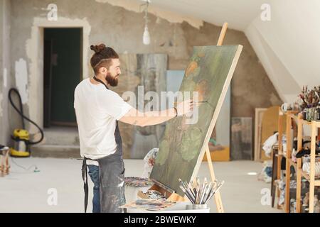 Portrait de l'artiste homme talentueux peint sur chevalet tout en travaillant dans un studio d'art spacieux éclairé par la lumière du soleil, espace de copie Banque D'Images