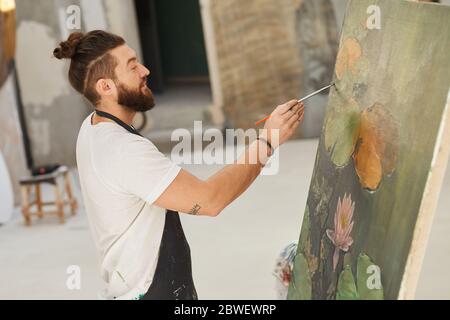 Portrait d'un artiste barbu talentueux, qui peint sur chevalet et souriait tout en appréciant le travail dans un studio d'art spacieux, espace de copie Banque D'Images