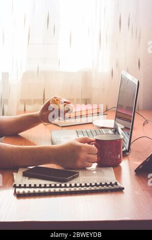 Homme travaillant à domicile. Utilisation de l'ordinateur et manger des beignets. Travail à distance ou éducation en ligne ou séjour à la maison. Banque D'Images