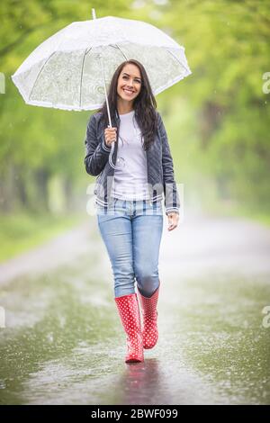 Une jeune femme heureuse marche avec un parapluie sous la pluie dans la nature, portant des bottes de pluie, un Jean, une veste et un t-shirt. Banque D'Images