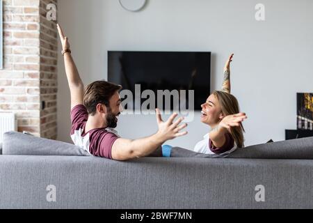 Un jeune couple regarde la télévision avec des mains levées pour célébrer la victoire assise sur un canapé Banque D'Images