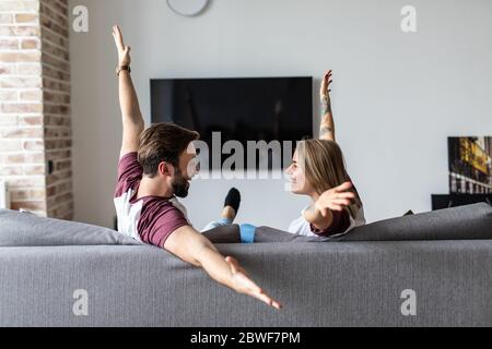 Un jeune couple regarde la télévision avec des mains levées pour célébrer la victoire assise sur un canapé Banque D'Images