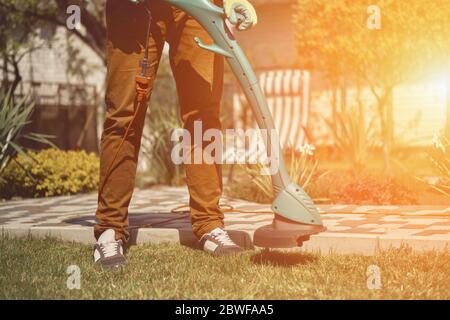 Guy dans les vêtements décontractés et les gants colorés est de couper l'herbe verte avec mini tondeuse électrique professionnelle dans le jardin de sa maison de campagne. Gros plan Banque D'Images