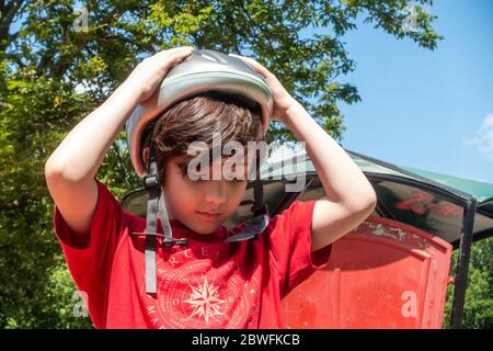 Un jeune enfant qui met un casque de vélo dans le parc attache une boucle sur une mentonnière. Banque D'Images