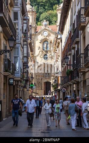 Touristes et habitants se baladant le long de Kale Nagusia à San Sebastian, Espagne avec la basilique ensoleillée de Saint Marie du Chorus en arrière-plan. Banque D'Images