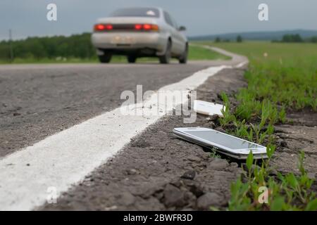 un téléphone portable cassé jeté est couché sur l'asphalte contre le fond d'une voiture qui passe Banque D'Images