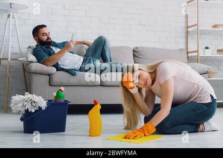 Sexisme et manque de respect pour la femme. L'homme se trouve sur le canapé et tape au téléphone, la triste femme se clins à la tête et lave les sols Banque D'Images