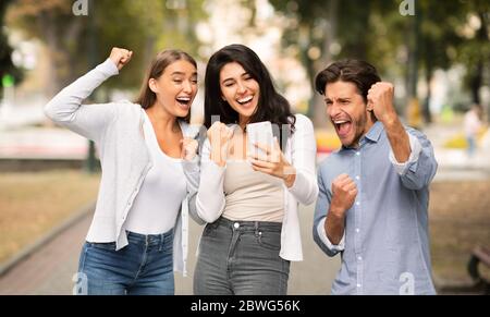 Trois amis joyeux avec des Fistes qui secouent leur téléphone cellulaire et célèbrent la victoire en plein air Banque D'Images