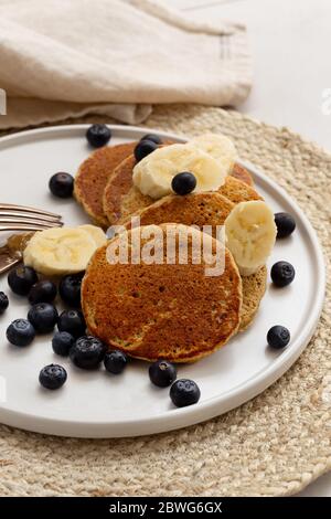 Image verticale des crêpes de flocons d'avoine à la banane servies avec les tranches de bananes et de bleuets sur la plaque ronde en céramique blanche placée sur le napperon de jute Banque D'Images