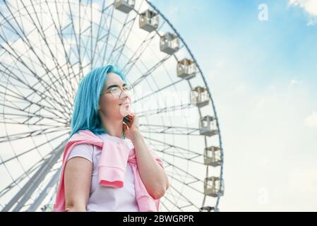 Portrait de jeune femme avec cheveux bleus et lunettes de soleil élégantes avec ciel nuageux et roulette de ferris en arrière-plan Banque D'Images