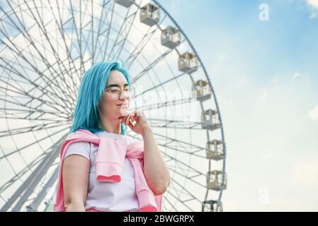 Portrait de jeune femme avec cheveux bleus et lunettes de soleil élégantes avec ciel nuageux et roulette de ferris en arrière-plan Banque D'Images