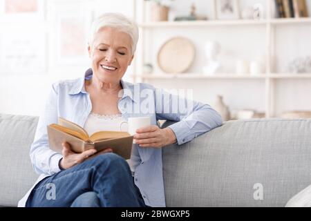 Temps de retraite. Femme âgée assise sur un canapé avec un livre et un café Banque D'Images