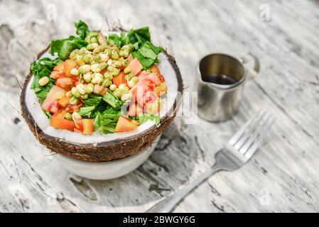 Salade de tomates, de légumes verts, de haricots mungs germés et de pulpe de noix de coco servie dans une demi-noix de coco sur fond de bois blanc Banque D'Images