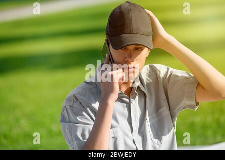 Jeune adolescent portant un chapeau à l'extérieur, sur fond d'herbe verte, qui chat sur son téléphone portable Banque D'Images