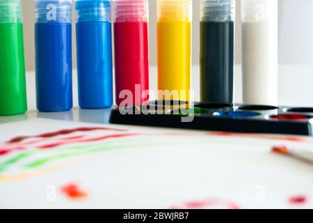 Papier blanc avec dessin d'enfant fait de peinture colorée avec plusieurs contenants de peinture acrylique et une tasse en plastique avec de l'eau et des brosses à l'intérieur Banque D'Images