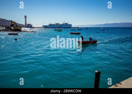 Aqaba, Jordanie - 2 février 2020 : deux pêcheurs reviennent à la terre en petit bateau à moteur après avoir pêché dans la mer Rouge. Grand bateau de croisière en arrière-plan. Ciel clair et sans nuages jour d'hiver. Cadre horizontal Banque D'Images