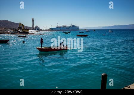 Aqaba, Jordanie - 2 février 2020 : deux pêcheurs reviennent à la terre en petit bateau à moteur après avoir pêché dans la mer Rouge. Grand bateau de croisière en arrière-plan. Ciel clair et sans nuages jour d'hiver. Cadre horizontal Banque D'Images