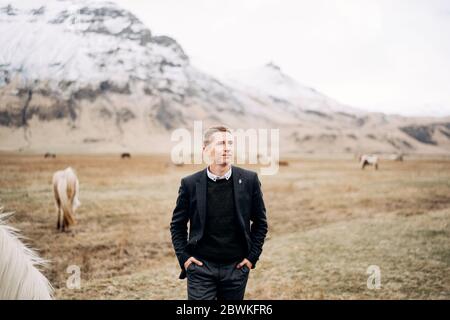 Un portrait épique de sexe masculin dans un champ d'herbe jaune, parmi les chevaux de pâturage, contre une montagne enneigée. Destination Islande mariage séance photo avec Banque D'Images