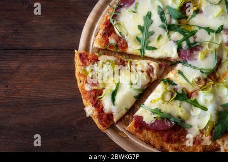 Partie d'une pizza à carb bas avec croûte de chou-fleur déchiqueté, surmontée de salami, roquette, fromage et poireau sur une table en bois rustique sombre, espace de copie, Banque D'Images