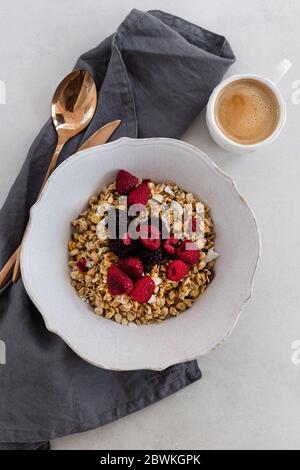 Café et granola aux baies dans un bol rustique blanc placé sur un plan de table gris clair. Banque D'Images