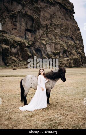 Une fille dans une robe blanche à côté d'un cheval blanc, avec des noirs. Jambes et manie. Dans un champ d'herbe sèche, sur fond de falaise. Destination Banque D'Images