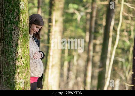 Une jeune femme enceinte souriante se tenant derrière un arbre touchant son ventre avec amour. Banque D'Images