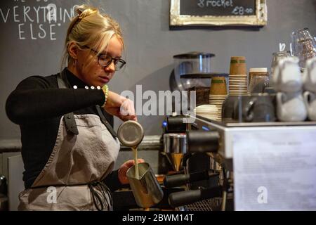 Femme blonde portant des verres et un tablier debout à la machine à espresso dans un café, versant du lait dans un pot métallique. Banque D'Images