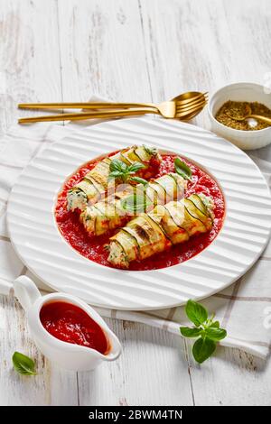 Courettes de courgettes farcies à la ricotta et aux épinards cuites dans une sauce tomate, servies sur une assiette avec une feuille de basilic fraîche sur un fond de bois blanc avec c Banque D'Images