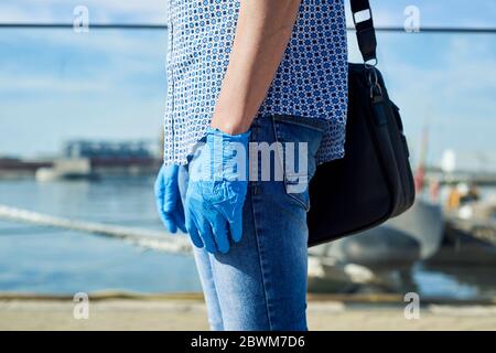 gros plan d'un jeune homme, portant des gants en latex bleu, une chemise et un jean, portant une mallette suspendue de son épaule, sur un port, en face de la mer Banque D'Images