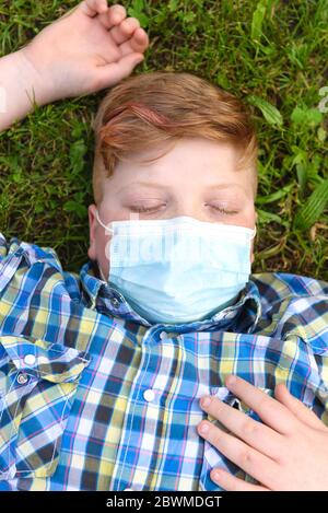 Portrait d'un jeune garçon avec masque chirurgical couché sur l'herbe dans le parc. Gros plan d'un petit garçon dans une chemise à carreaux se détendant sur l'herbe. Banque D'Images