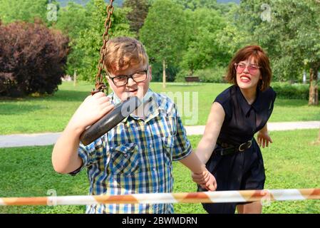 Un jeune garçon portant des lunettes et un masque chirurgical vêtu d'une chemise à carreaux joue avec une fille adulte devant un arou de ruban barrière rayé rouge et blanc Banque D'Images