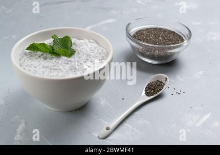 Le pudding de Chia avec des feuilles de menthe dans un bol blanc et un bol en verre avec des grains sur fond gris. Mise au point sélective. Un petit déjeuner sain se compose de yogu Banque D'Images