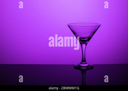 Gobelet en verre sans vin sur une jambe mince se tient sur une surface miroir. Rétroéclairage. Fond violet. Banque D'Images