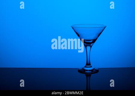 Gobelet en verre sans vin sur une jambe mince se tient sur une surface miroir. Rétroéclairage. Arrière-plan bleu. Banque D'Images