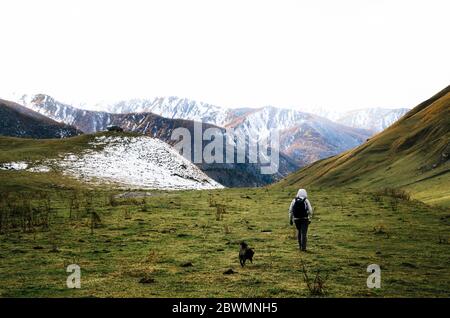 Femme voyageur avec chien marche sur la prairie verte à côté de la chaîne de montagnes qui couvrait avec des nuages gris. Vue arrière. Caucase, Géorgie Banque D'Images