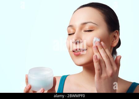 une jeune femme brune belle utilise un hydratant facial. isolé sur fond bleu Banque D'Images