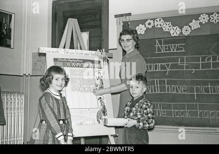 L'éducation aux Etats-Unis dans les années 1950 – dans une classe, un enseignant et deux élèves sont devant un tableau blanc avec un tableau sur l'hygiène personnelle. Les questions « vous vous lavez ? » et « vous vous brossez les dents ? » font partie de ce programme destiné aux élèves de première année de l'école élémentaire. Il y a des nouvelles écrites à la main sur le tableau noir derrière eux. Banque D'Images