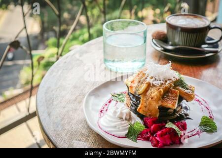Pile de crêpes végétaliennes décorées de banane, tranche de fruits de dragon, chocolat et crème de noix de coco avec café et eau sur la table Banque D'Images