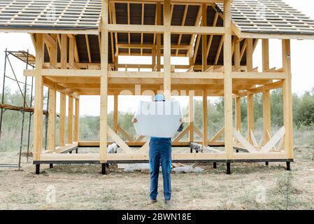 Constructeur en combinaison bleue et chapeau de dur avec des plans sur le chantier de construction, bâtiment en bois cadre maison Banque D'Images